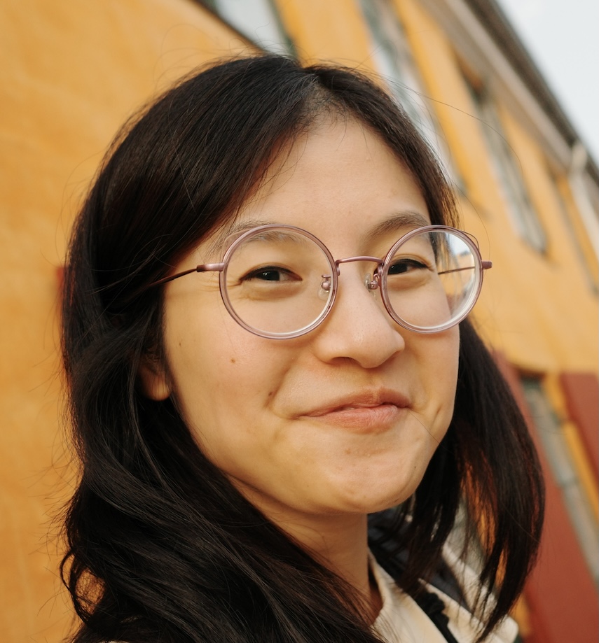 Wide and close portrait of an East Asian woman with long dark hair, in round purple wire-rimmed glasses, smiling with lips closed and head tilted toward the camera. Her face fills the center and the frame tips off-level at a Dutch angle. Behind, an ochre plaster wall runs diagonally into the distance, punctuated by dark red shutters.
